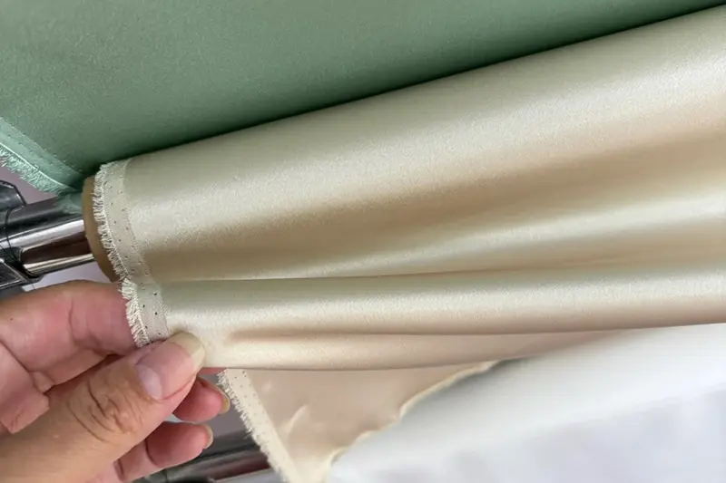 Hand lifting the edge of an ivory fabric roll to reveal its sheen