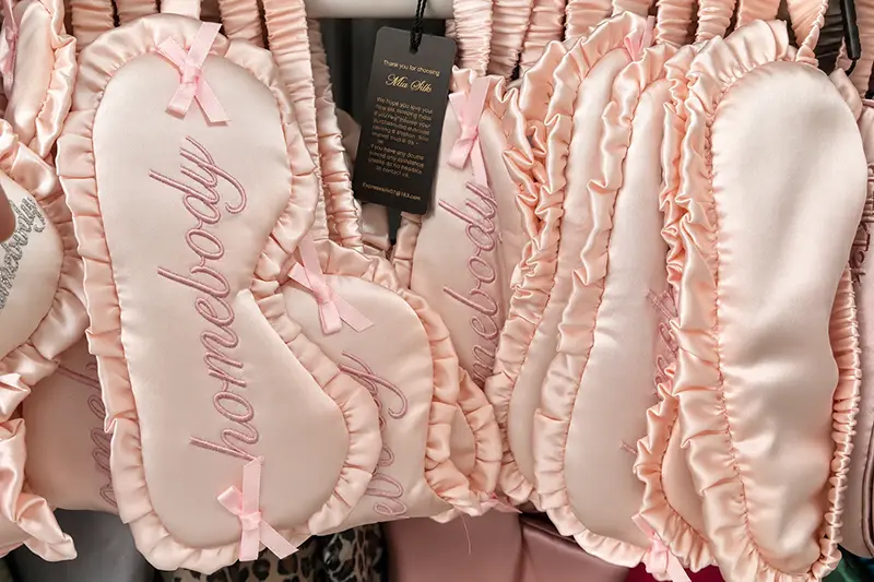 Pink ruffle-edged silk sleep masks with embroidered lettering hanging on display