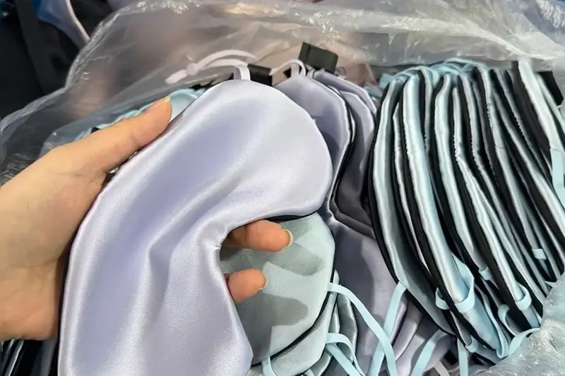 Hand lifting a pale sleep mask beside stacked finished masks